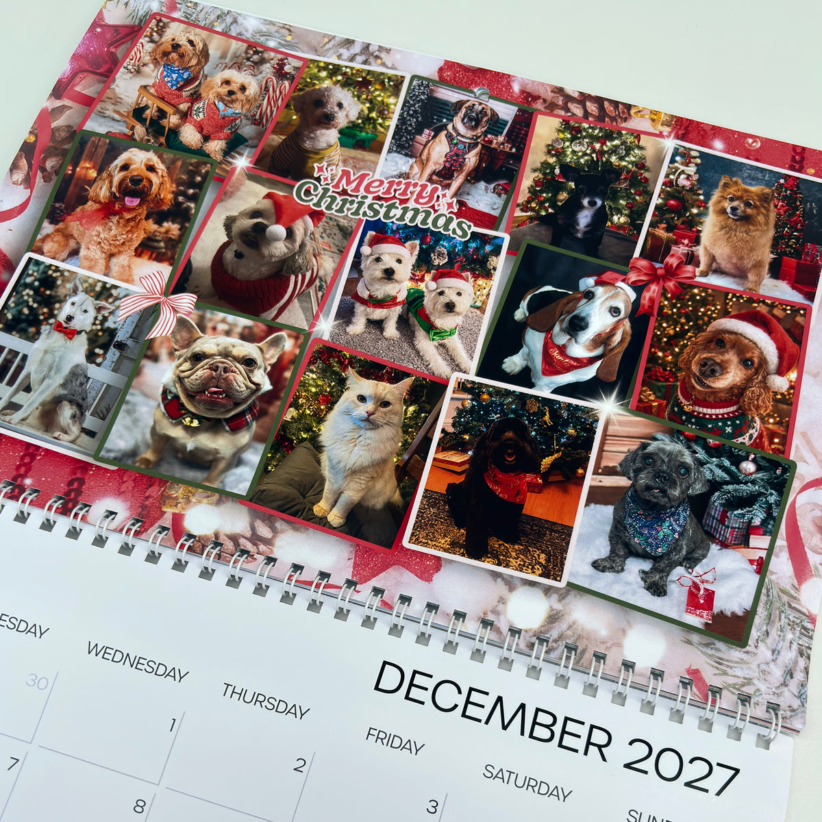 A spiral-bound 2027 calendar page featuring a collage of holiday dog photos with Christmas decorations, bows, and a 'Merry Christmas' banner, with the December 2027 date visible at the bottom.