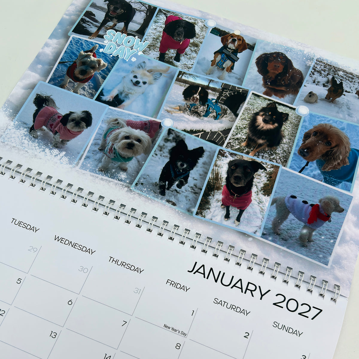 A spiral-bound January 2027 wall calendar page featuring a collage of dog photos in the snow under the words 'SNOWY DAY!'.