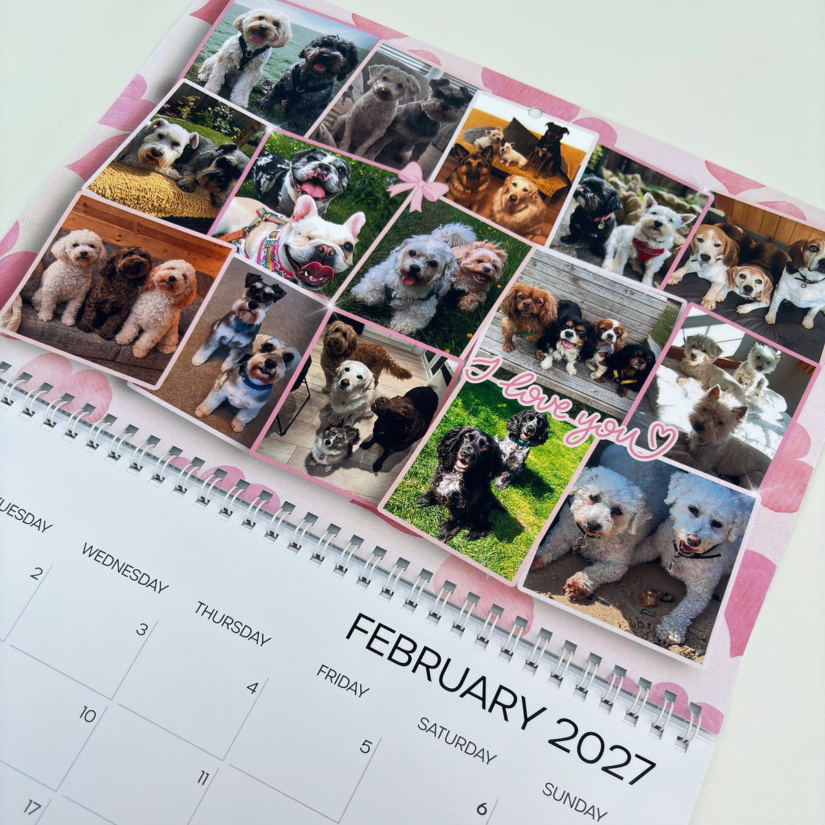 A spiral-bound 2027 desk calendar page featuring a collage of various dog photos, pink floral borders, and the text 'I love you' with a heart. The calendar is photographed on a clean white surface.